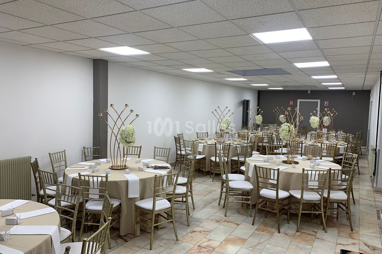 Salle de réception décorée avec des tables rondes, nappes beige, chaises dorées et centres de table floraux.