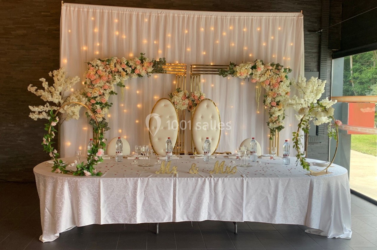 Table décorée pour un mariage avec nappes blanches, fleurs, chaises élégantes et arrière-plan lumineux.