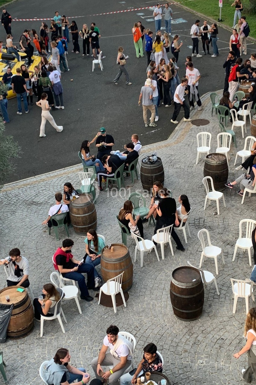 Groupe de personnes assises et debout autour de tonneaux et de chaises sur une place pavée, ambiance conviviale en extérieur.