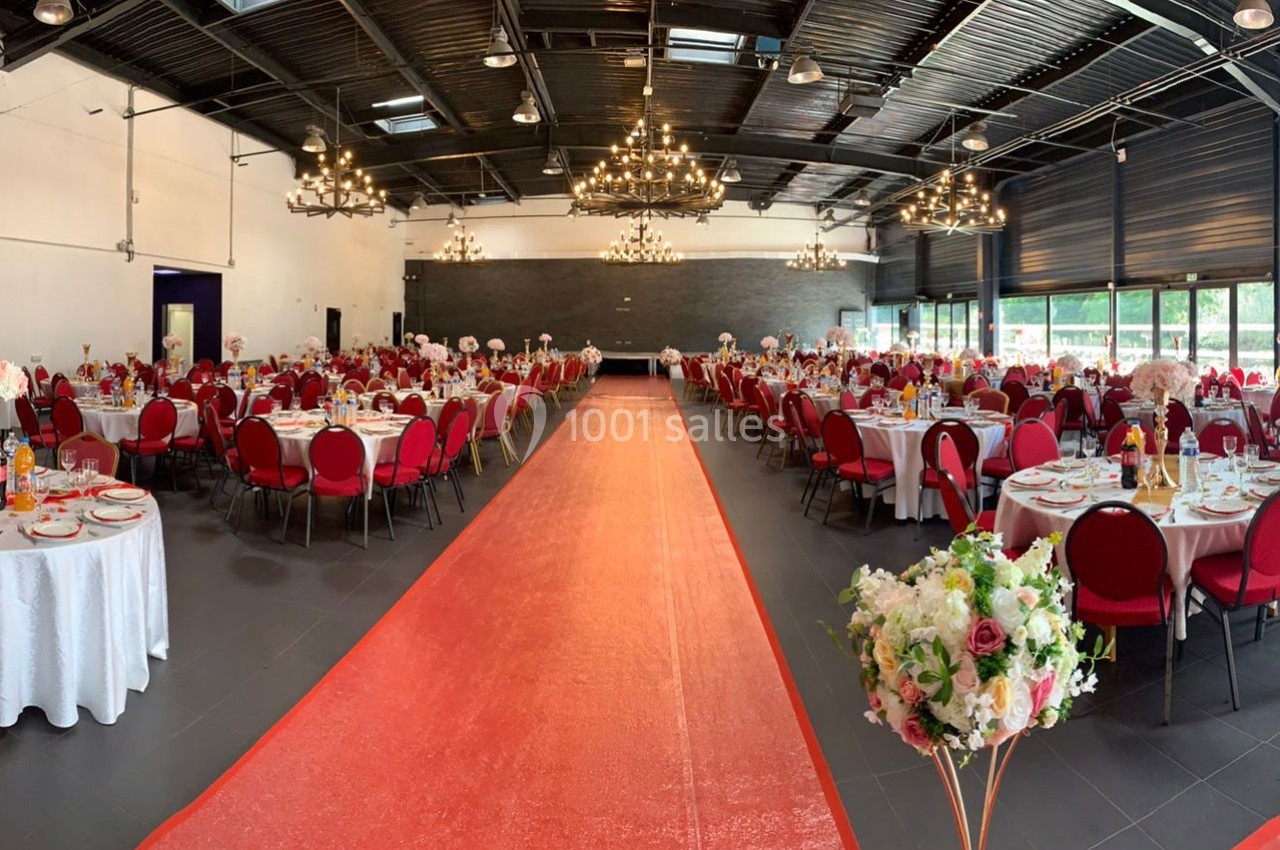 Salle de réception décorée avec des tables rondes dressées, chaises rouges et un tapis rouge central.