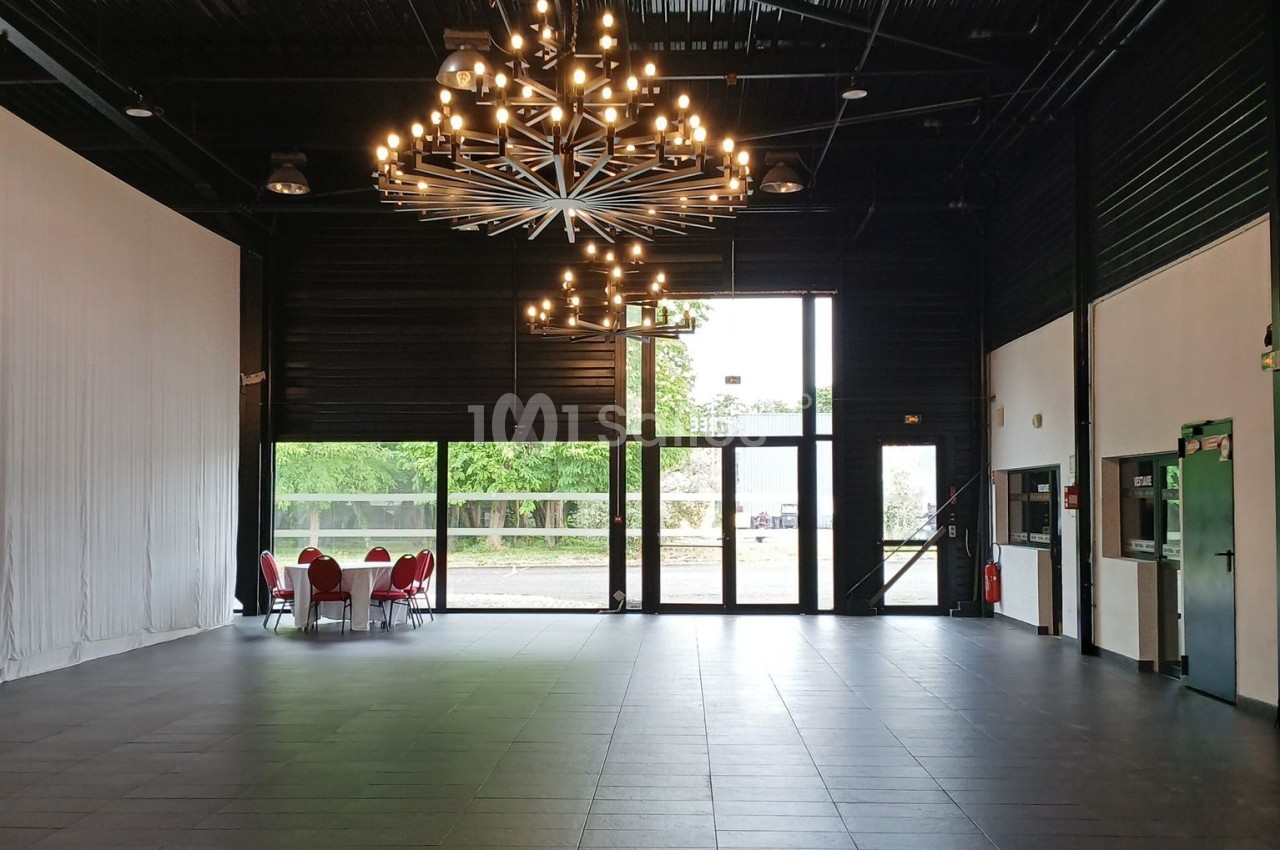 Salle spacieuse avec un sol carrelé, un grand lustre central, des baies vitrées et des chaises rouges près d'une table.
