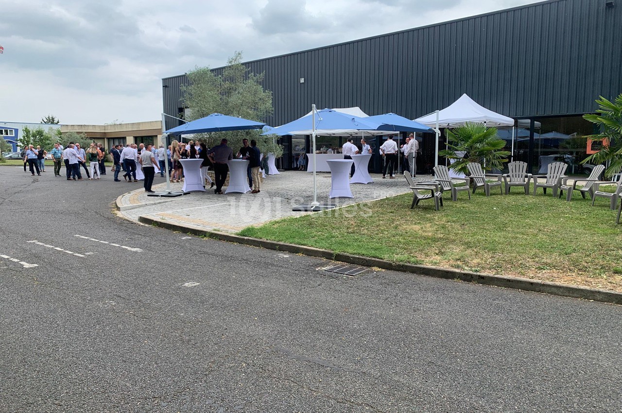 Personnes rassemblées autour de tables sous des tentes blanches et bleues dans une cour extérieure près d'un bâtiment.