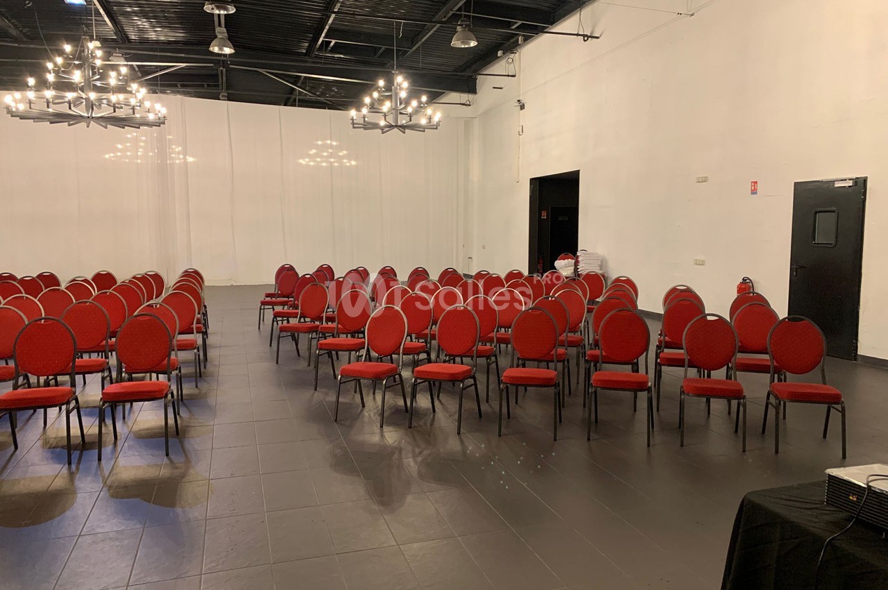 Salle de conférence avec rangées de chaises rouges alignées, éclairée par des lustres suspendus au plafond noir.