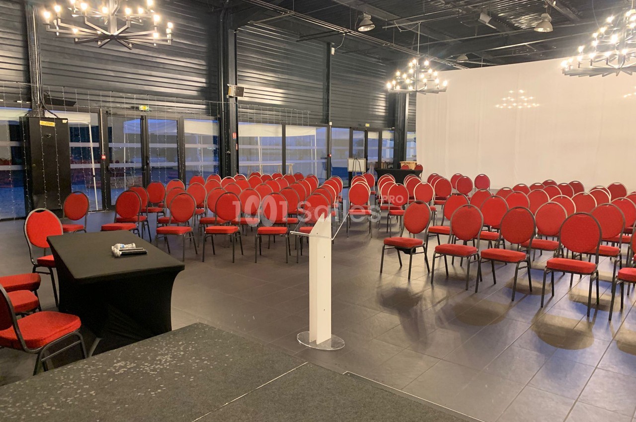 Salle de conférence avec des rangées de chaises rouges disposées face à un pupitre et une table noire.