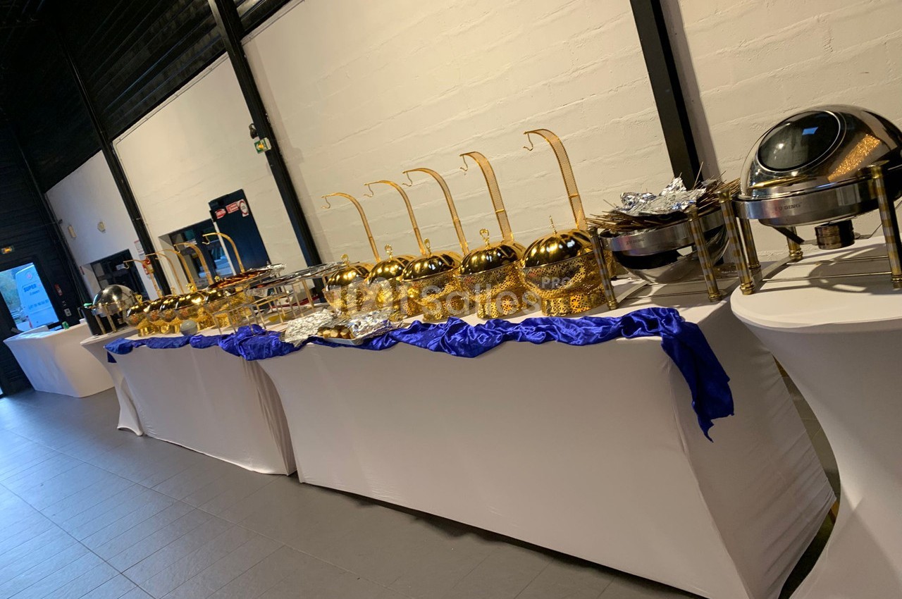 Buffet avec plats chauds présentés dans des récipients dorés et argentés sur des tables drapées de nappes blanches.
