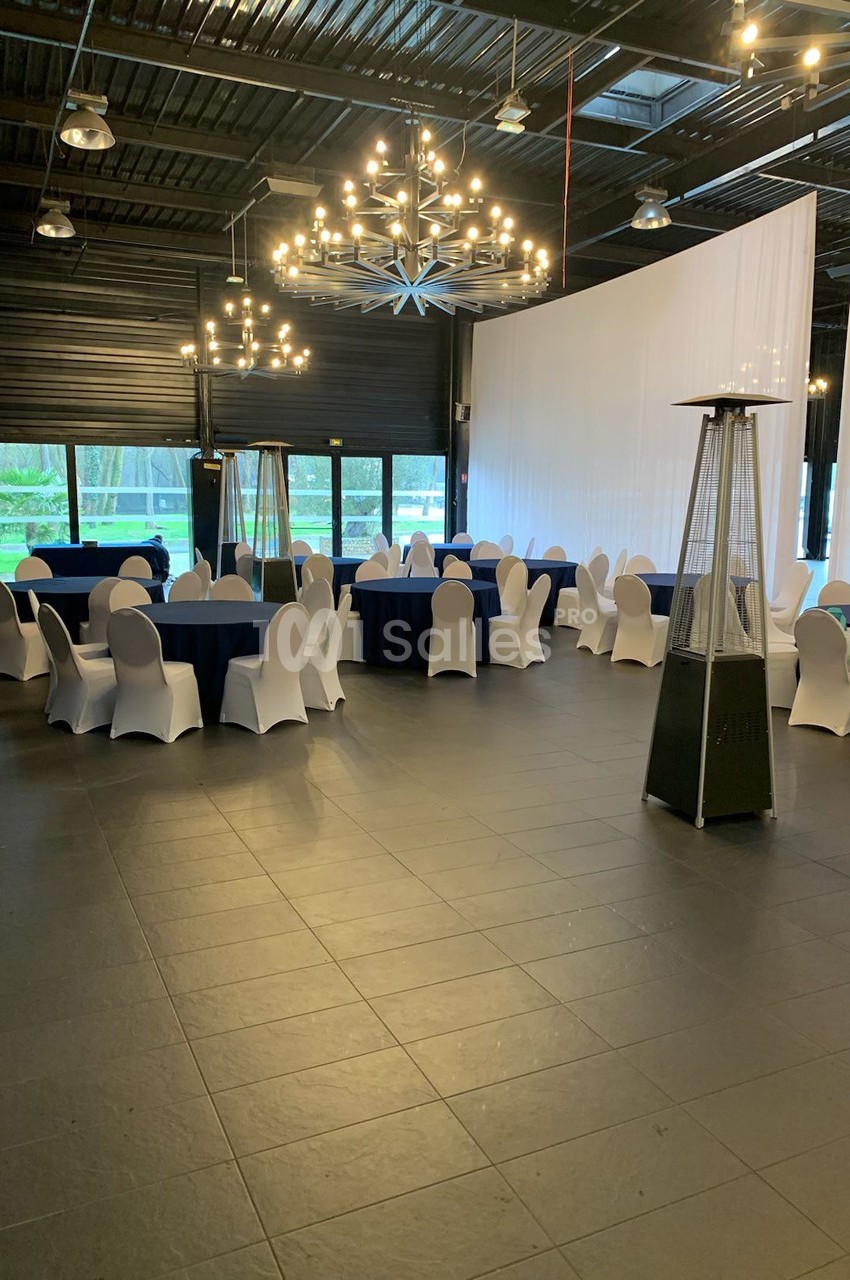 Salle de réception avec tables rondes dressées de nappes bleues, chaises blanches et grands lustres suspendus.