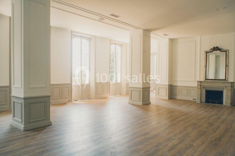 Salle lumineuse avec parquet en bois, murs blancs ornés de moulures, cheminée et grands miroirs près des fenêtres.