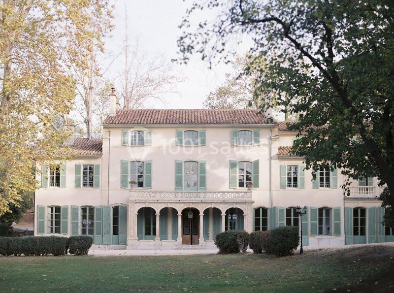 Grande maison provençale avec volets verts, façade claire et jardin arboré sous une lumière douce.
