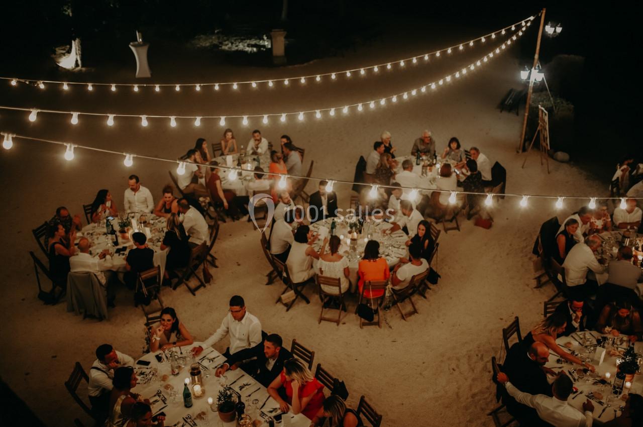Vue aérienne d'un dîner en plein air avec des invités assis à des tables rondes sous des guirlandes lumineuses.