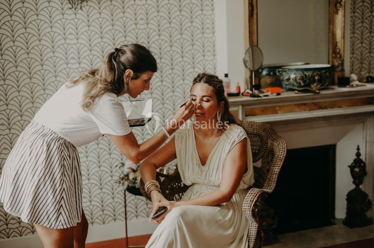 Une femme maquille une autre femme assise dans un fauteuil, dans une pièce au décor élégant avec une cheminée.