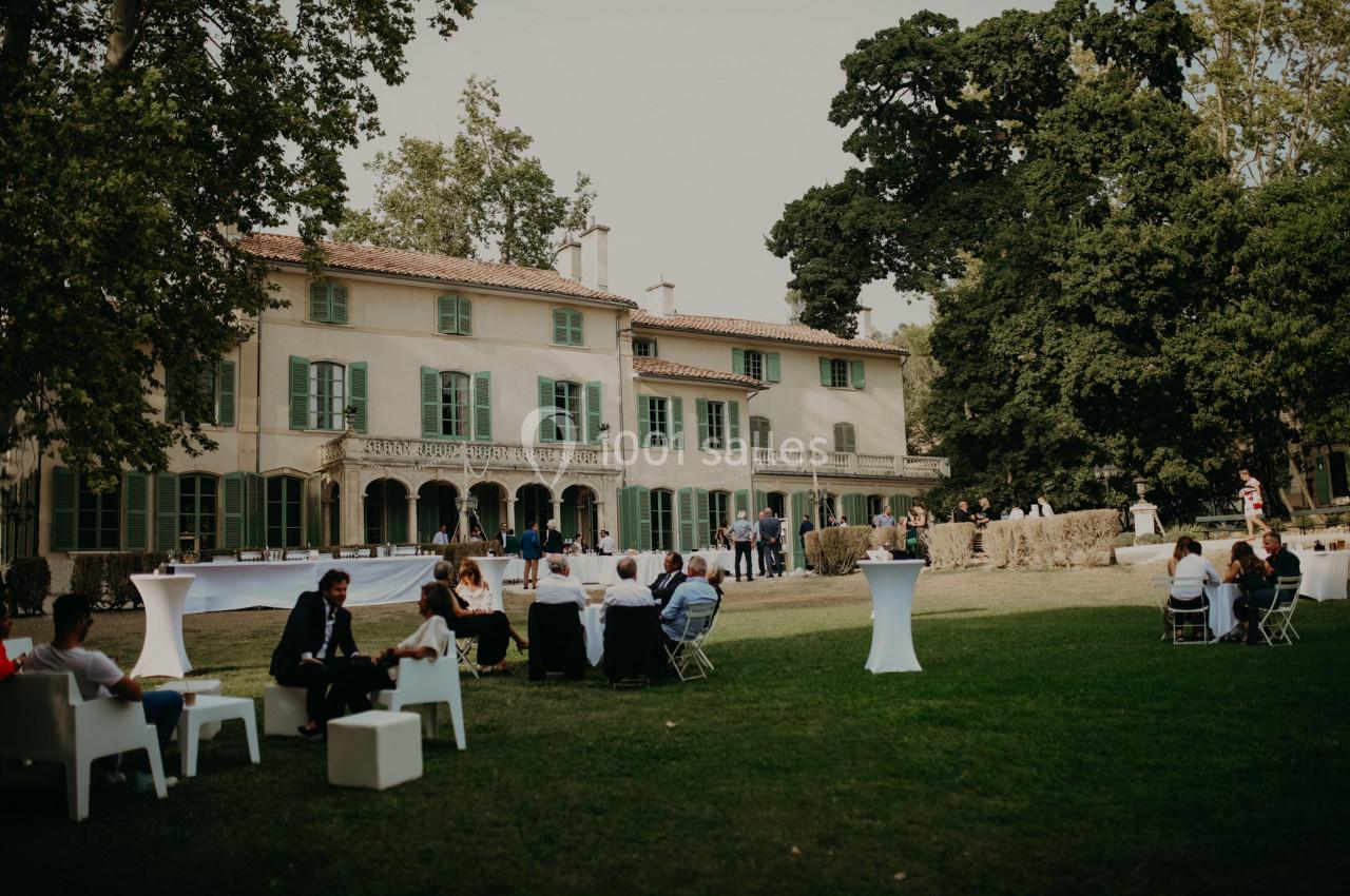 Personnes assises et debout dans un jardin devant une grande maison ancienne entourée d'arbres.