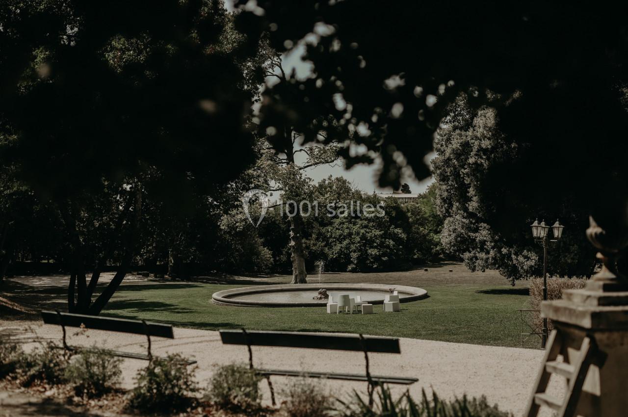 Bancs en bois face à une fontaine circulaire dans un parc arboré, sous une lumière tamisée.