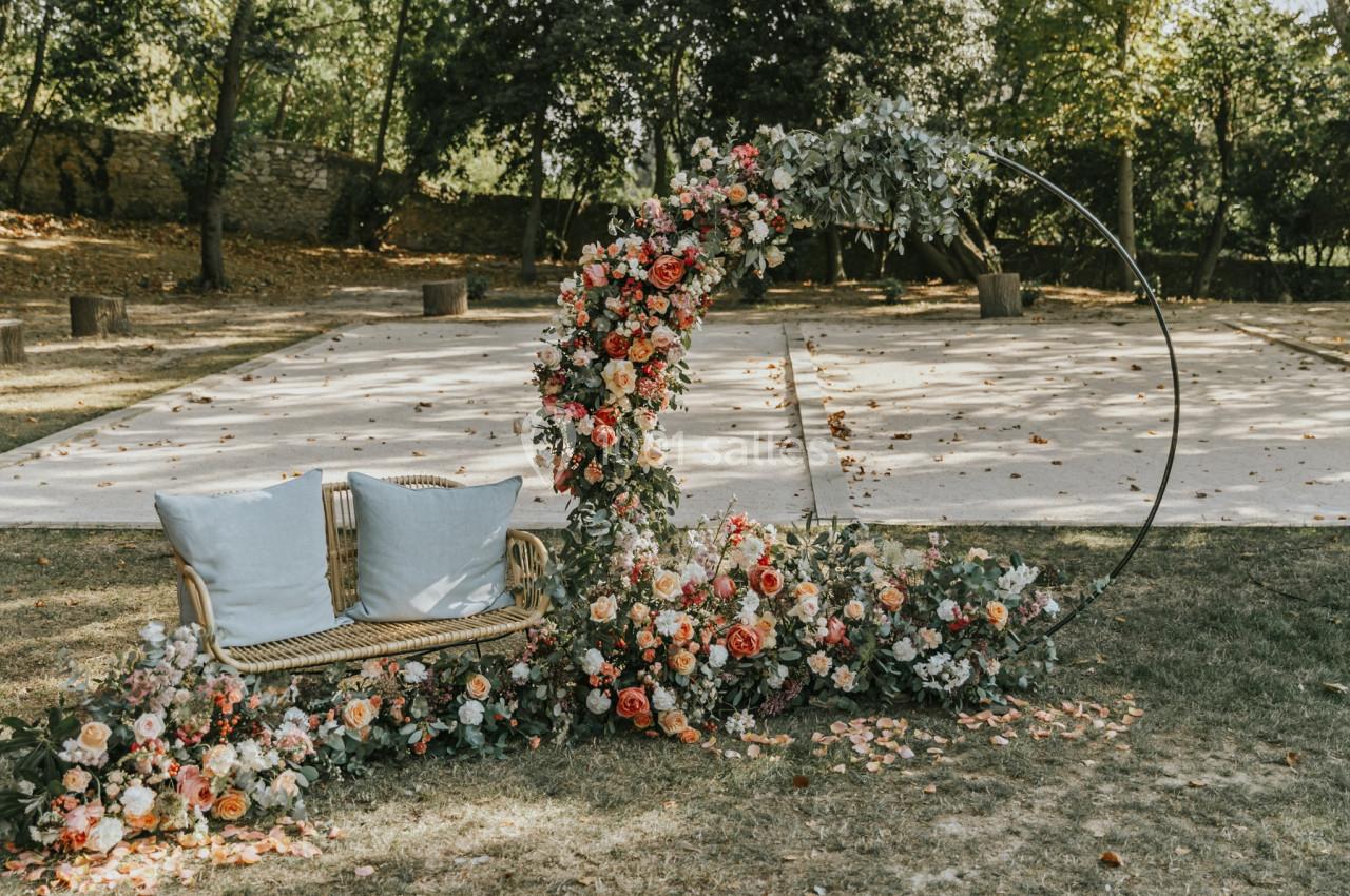 Arche circulaire décorée de fleurs colorées et banc en rotin avec coussins, installés dans un jardin ombragé.