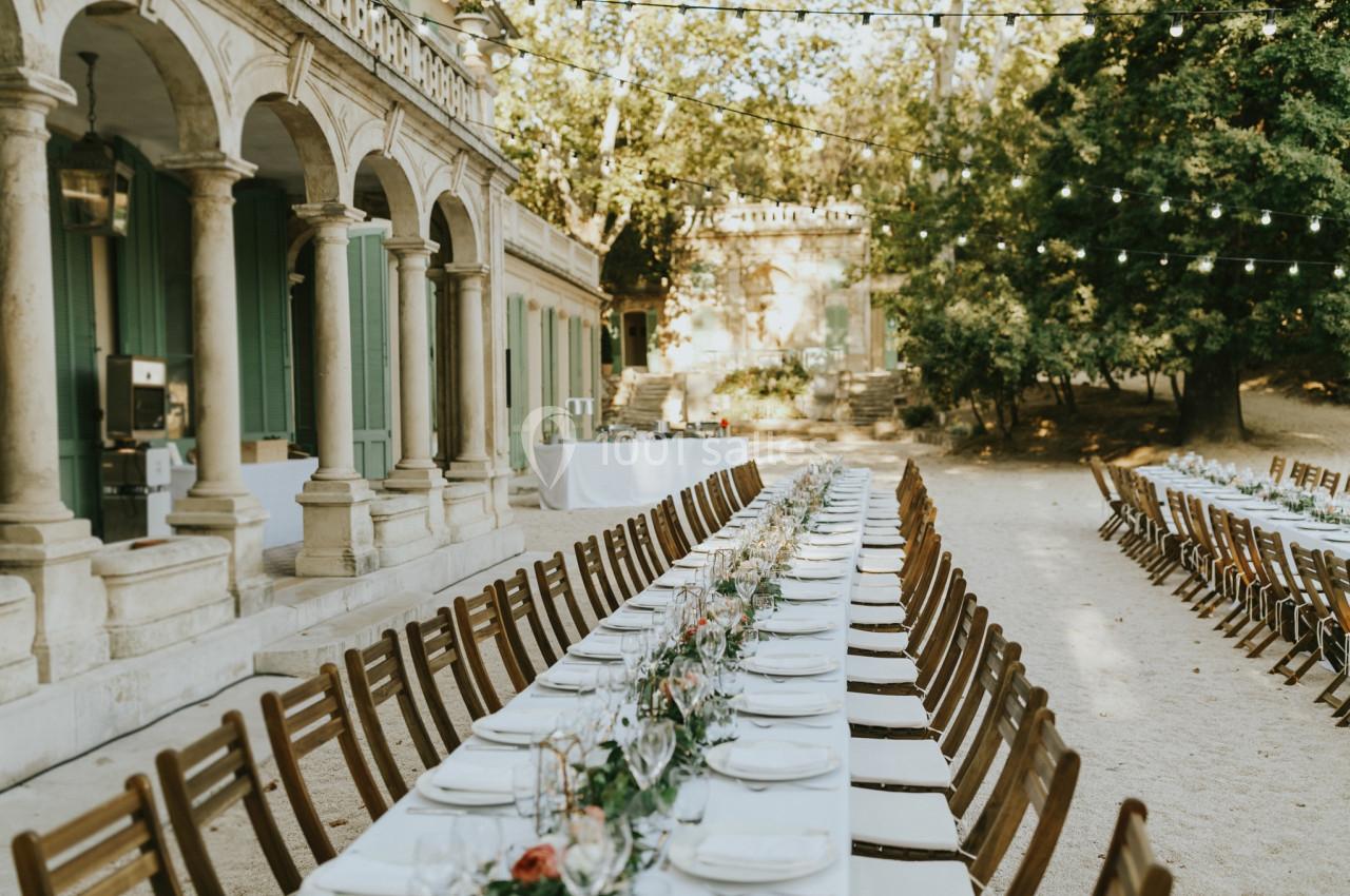Tables longues décorées pour un repas en extérieur, disposées sous des guirlandes lumineuses près d'un bâtiment ancien.