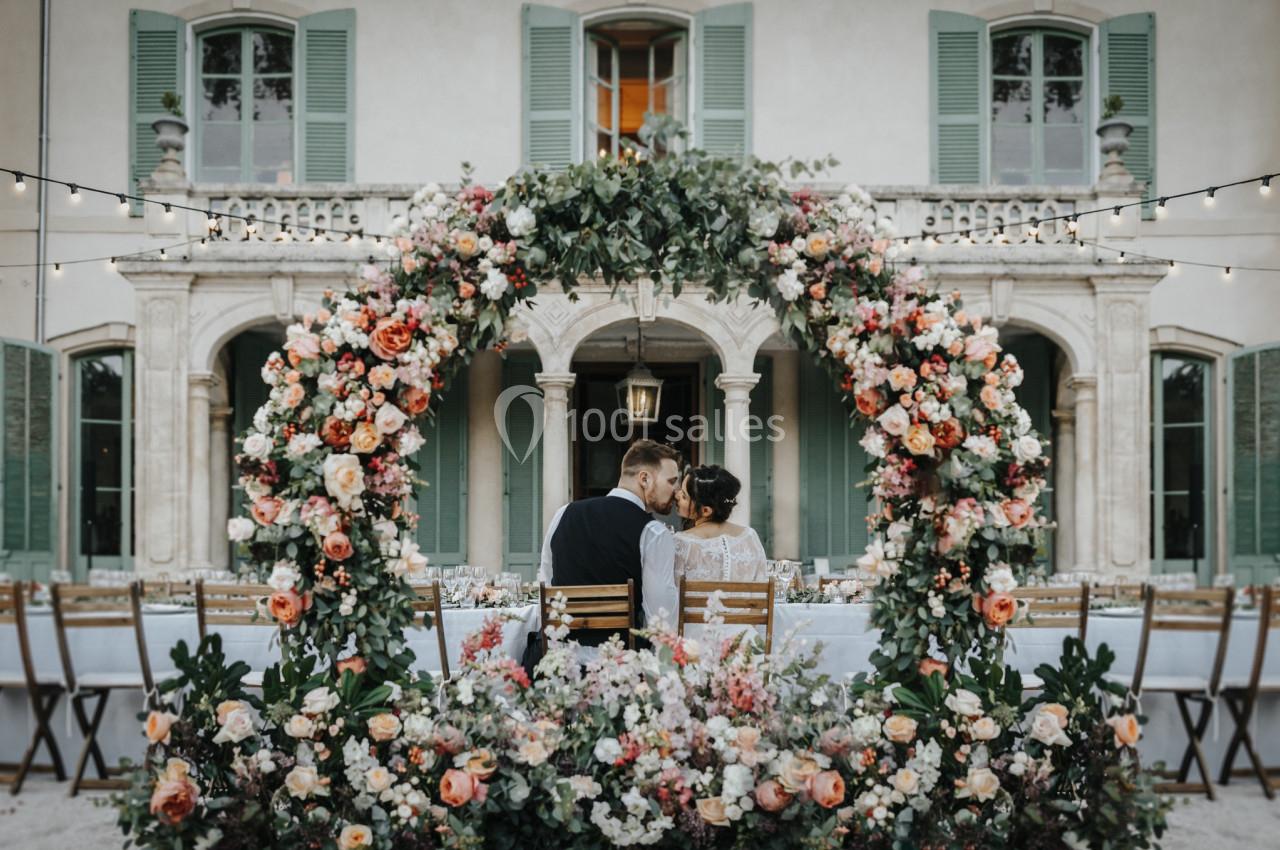 Un couple assis dos à une arche fleurie, face à une table décorée, devant une grande bâtisse aux volets verts.