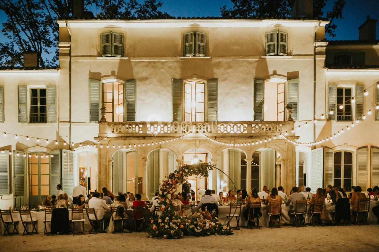 Dîner en plein air devant une grande bâtisse illuminée, avec des invités assis à une longue table décorée de fleurs.