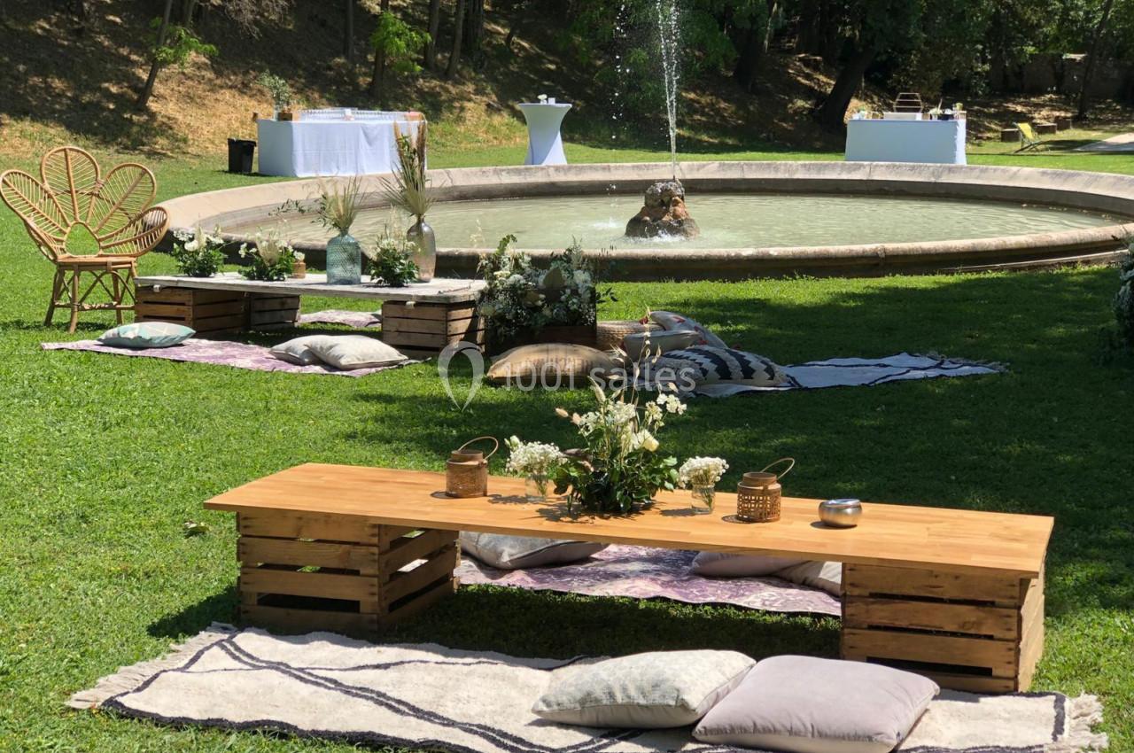 Décoration champêtre avec banc en bois, coussins et fontaine en arrière-plan dans un jardin verdoyant.