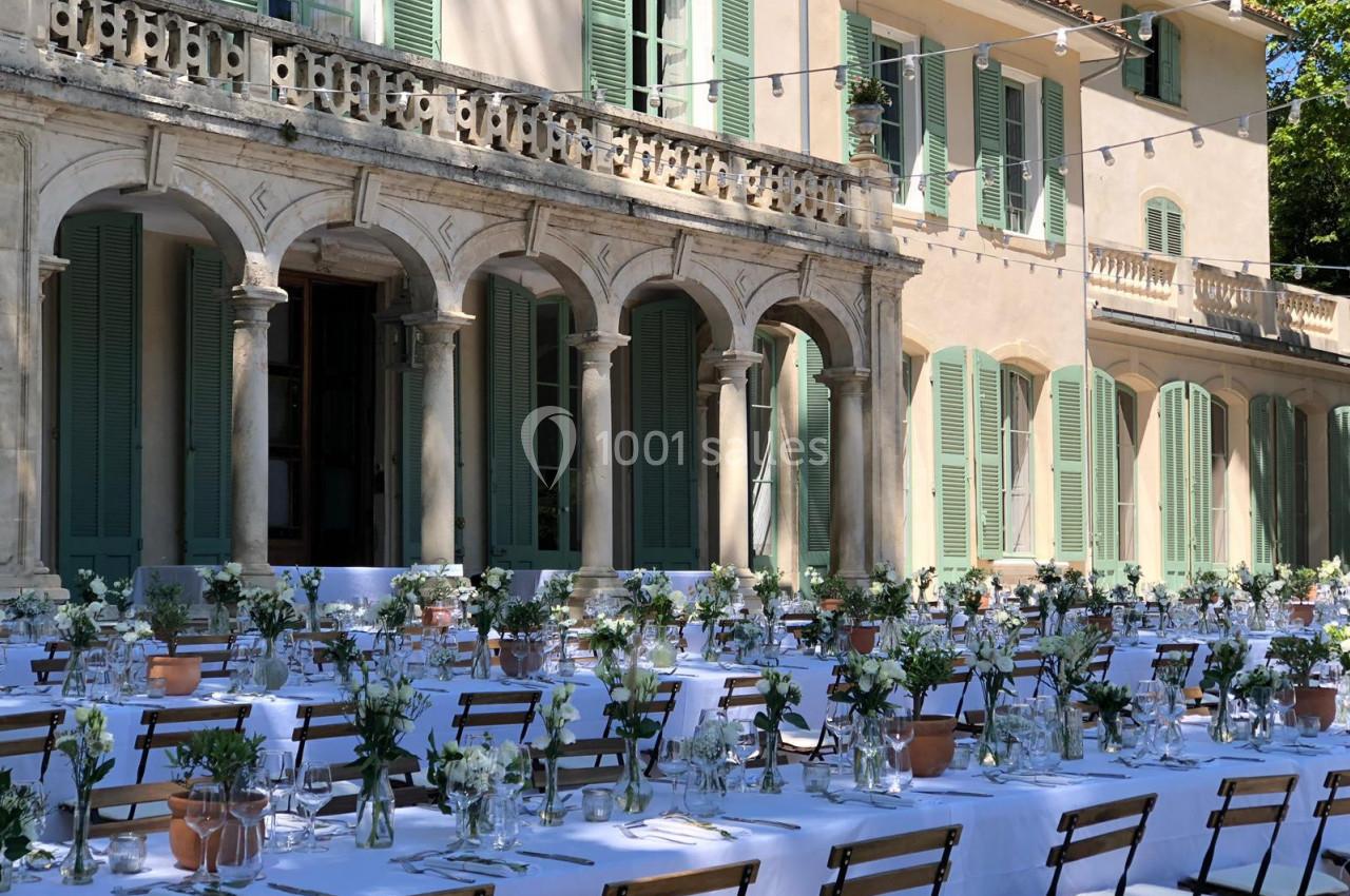 Tables dressées pour un repas en extérieur devant une grande bâtisse aux volets verts et à l'architecture classique.
