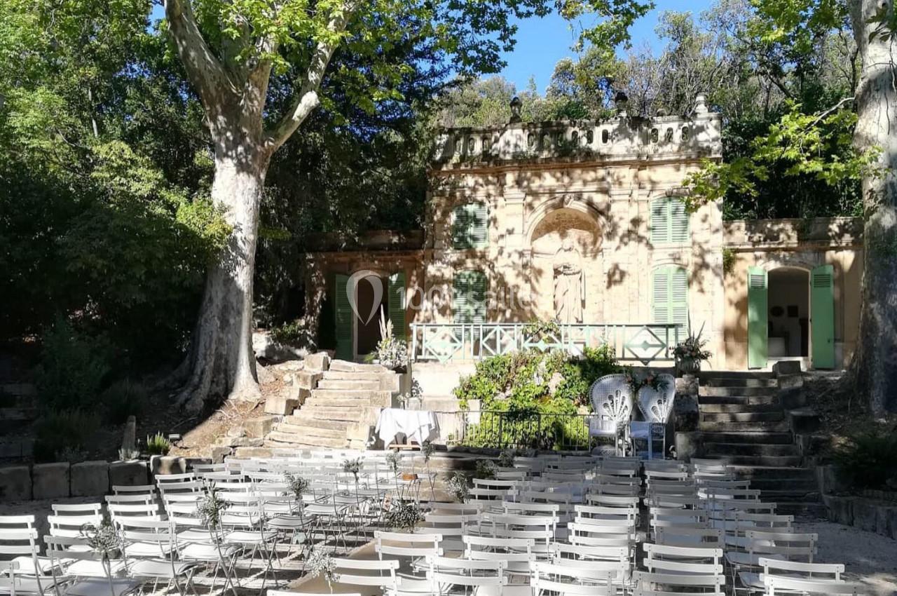 Chaises blanches alignées en extérieur devant une petite bâtisse ancienne entourée d'arbres et de verdure.