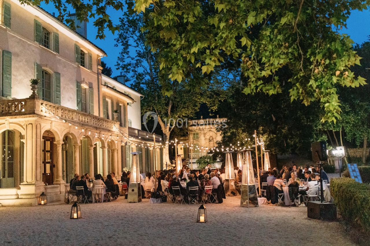 Dîner en plein air sous des guirlandes lumineuses devant un bâtiment ancien entouré d'arbres.