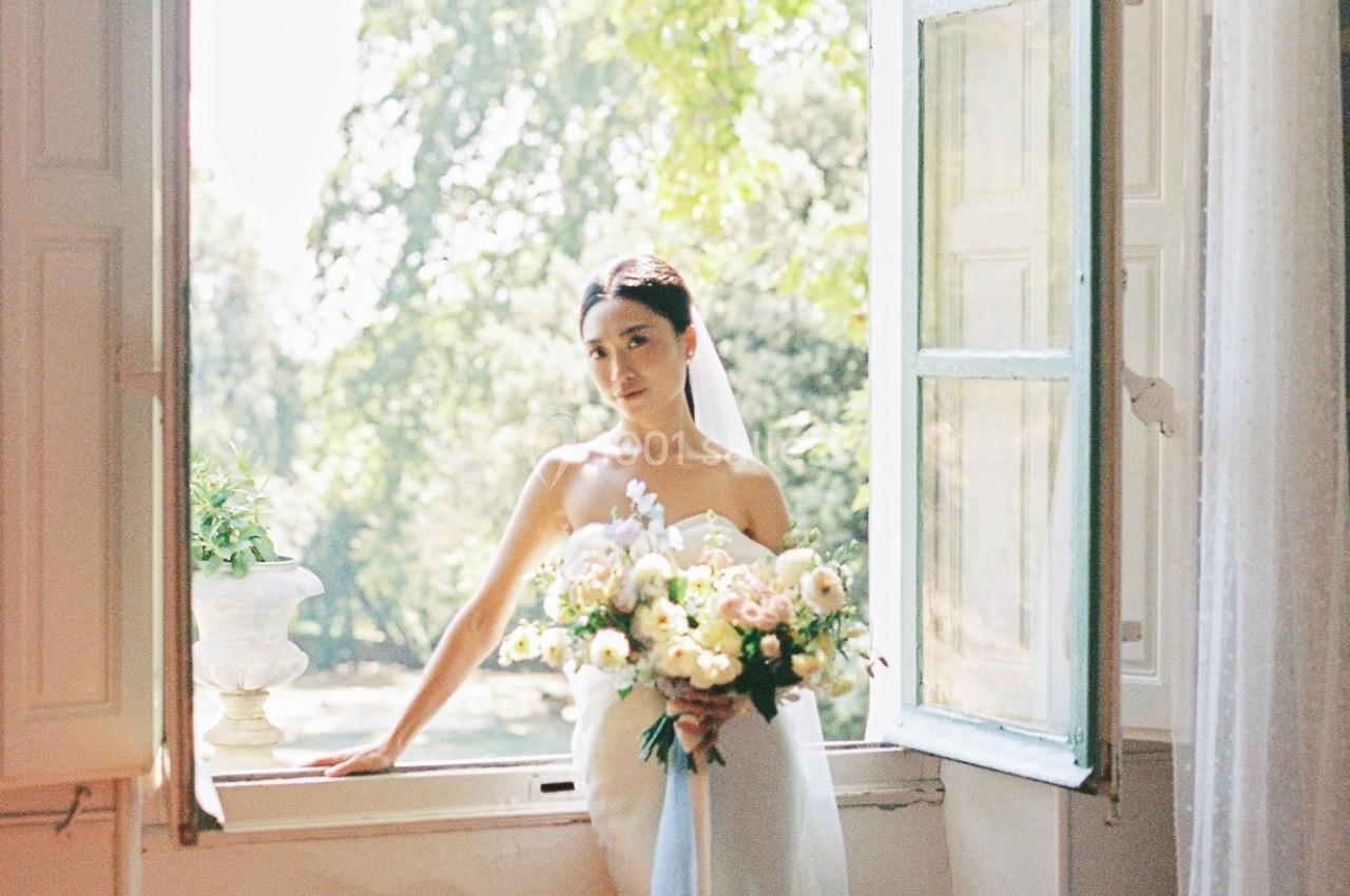 Une mariée en robe blanche tient un bouquet de fleurs devant une fenêtre ouverte avec vue sur des arbres verdoyants.