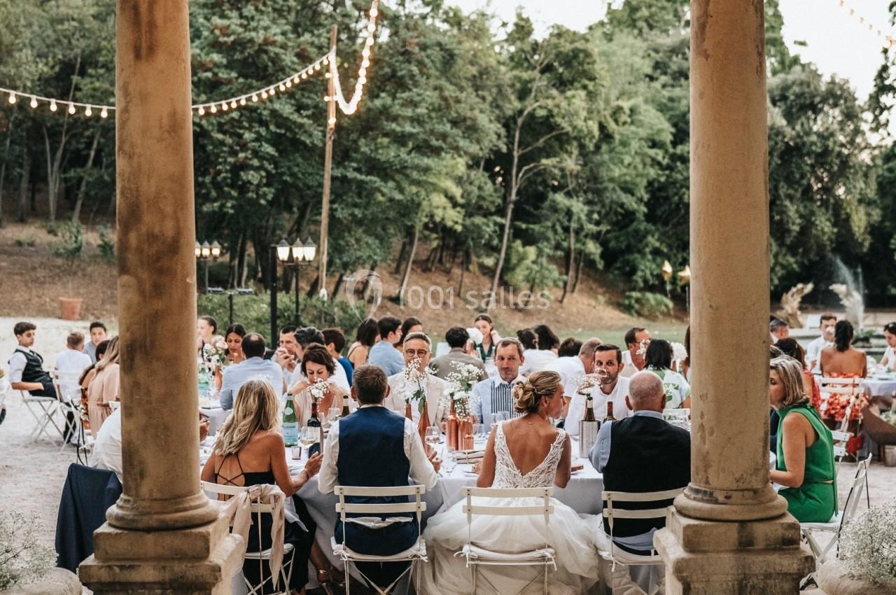Des invités assis à des tables décorées sous des guirlandes lumineuses dans une cour extérieure entourée d'arbres.