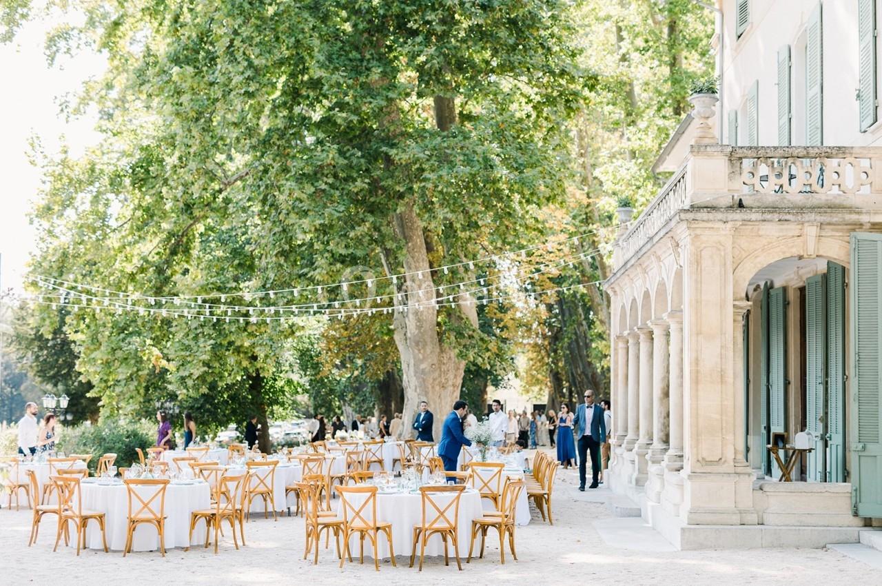 Tables et chaises en bois disposées en extérieur sous des guirlandes lumineuses près d'un bâtiment ancien entouré d'arbres.