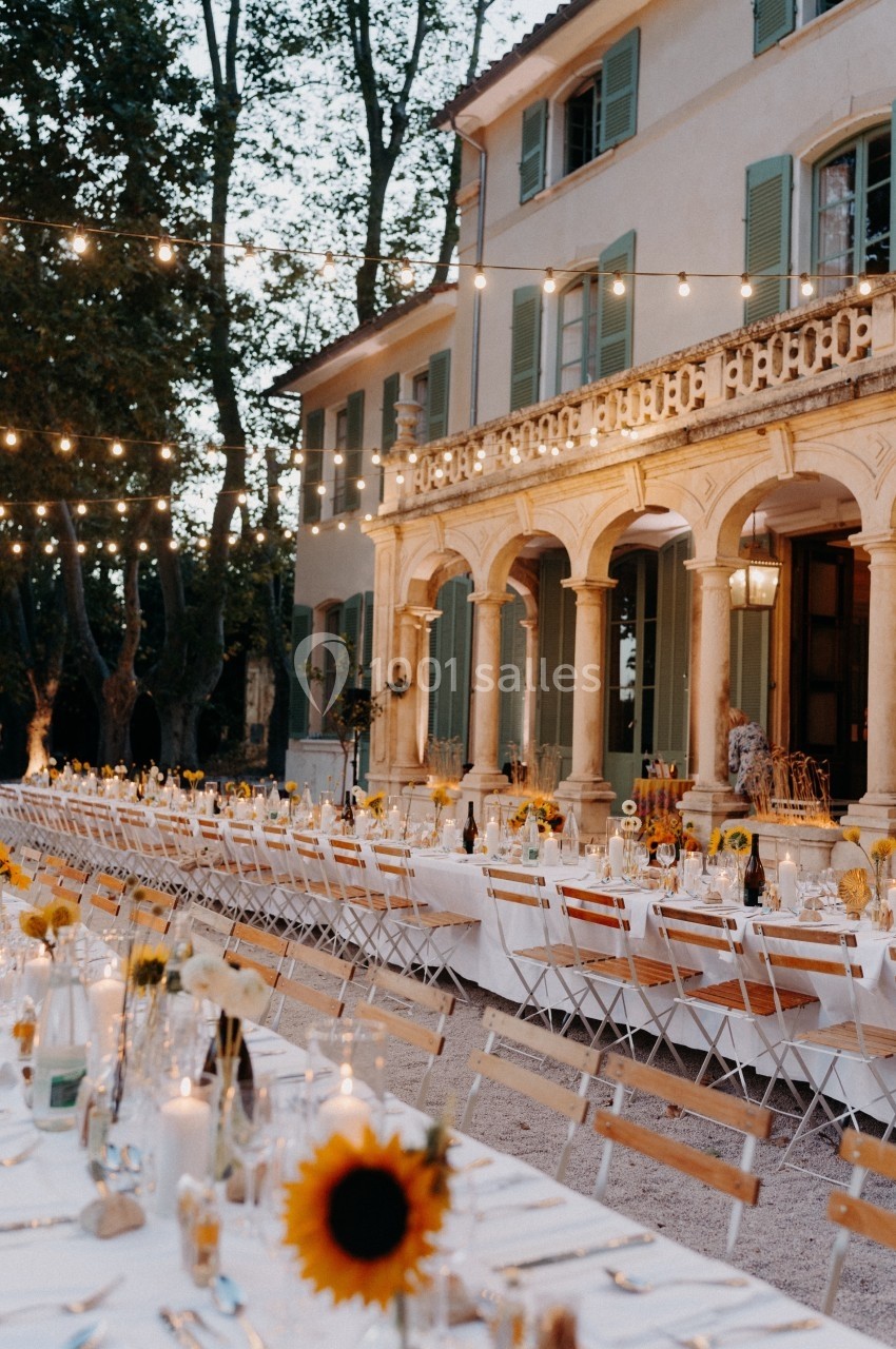 Grande table dressée en extérieur avec des guirlandes lumineuses, devant une maison élégante entourée d'arbres.