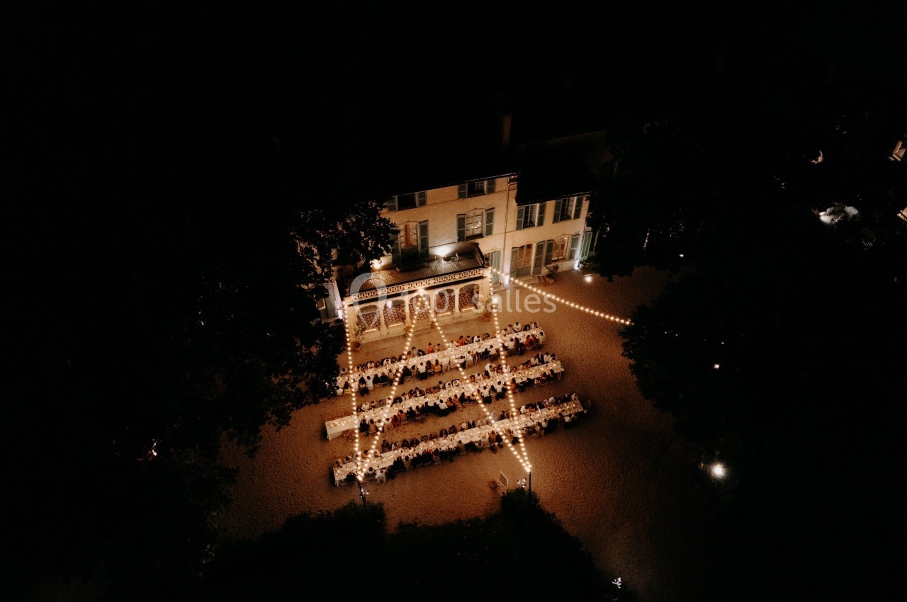 Vue aérienne nocturne d'une réception en extérieur, avec des tables alignées sous des guirlandes lumineuses près d'un…