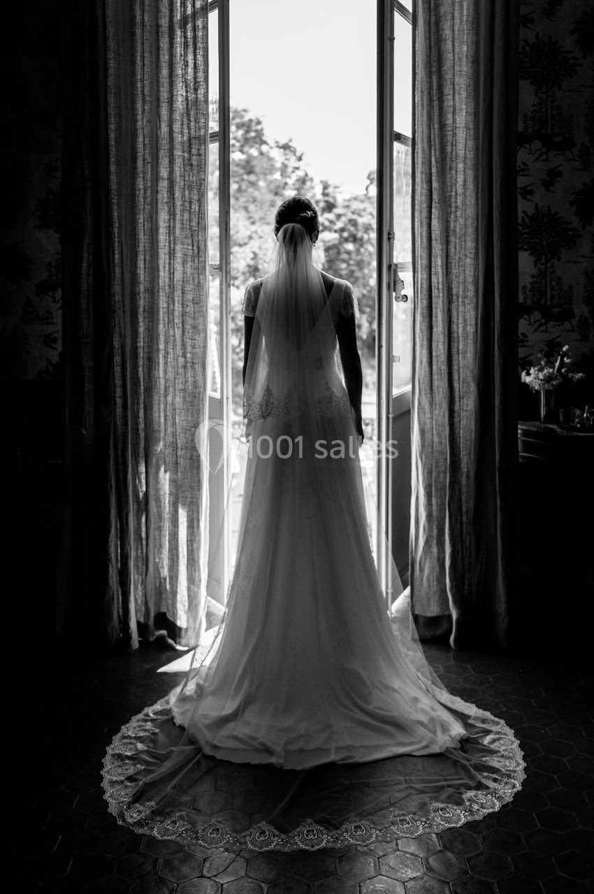 Une mariée en robe blanche et voile vue de dos, debout devant une porte-fenêtre ouverte sur un extérieur lumineux.