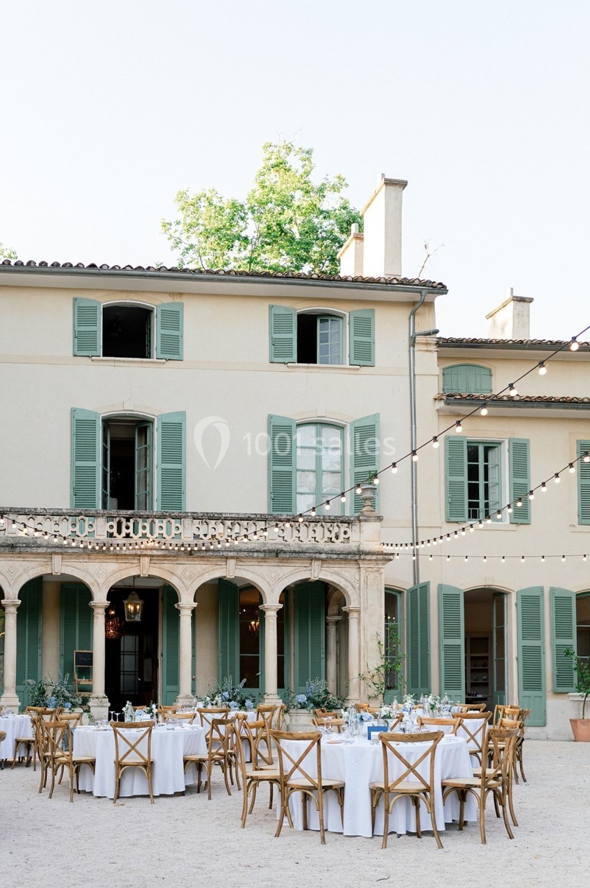 Tables dressées pour un repas en plein air devant une grande maison aux volets verts, éclairée par des guirlandes lumineuses.