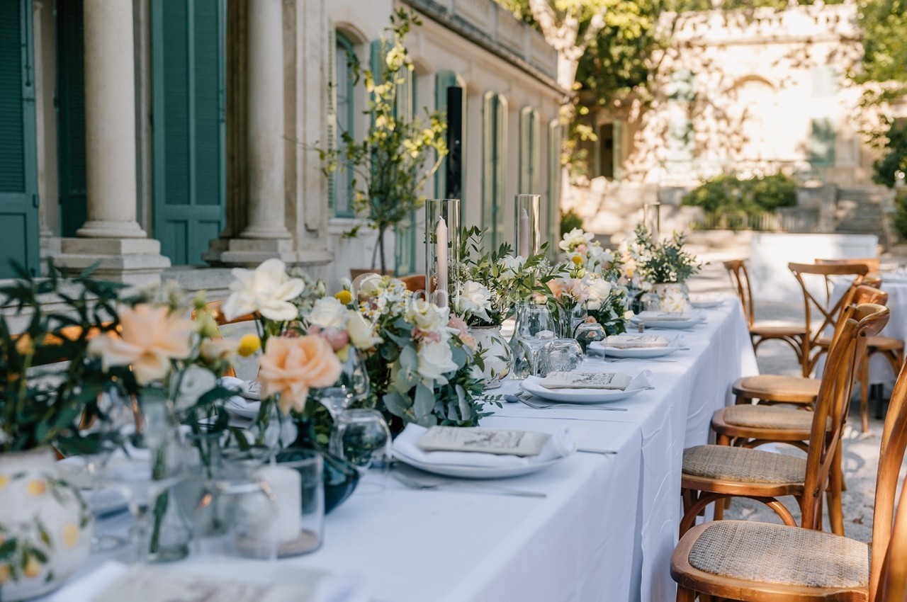 Table dressée en extérieur avec nappes blanches, fleurs pastel et chaises en bois devant un bâtiment ancien.