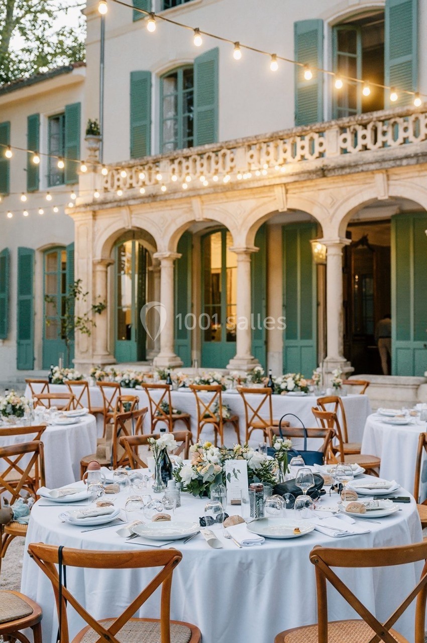 Tables de réception élégamment dressées en extérieur, devant une bâtisse aux volets verts, éclairée par des guirlandes…