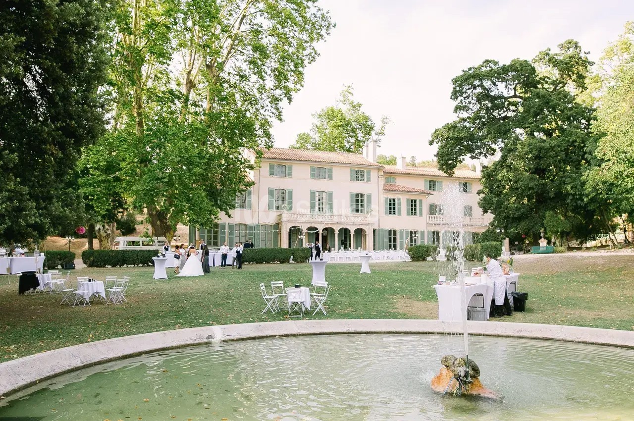 Jardin avec fontaine, tables dressées et invités devant une grande maison de style provençal entourée d'arbres.