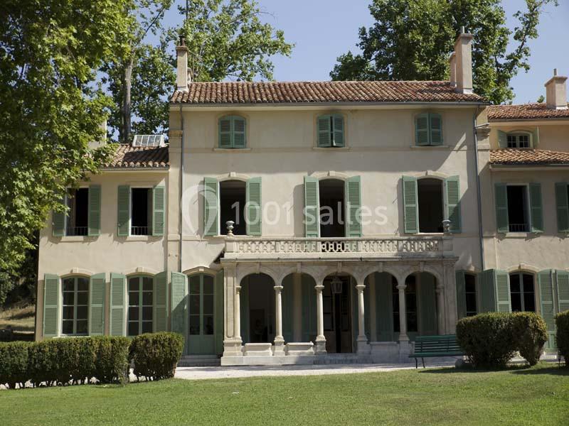 Façade d'une maison ancienne avec volets verts, balcon central et jardin arboré au premier plan.