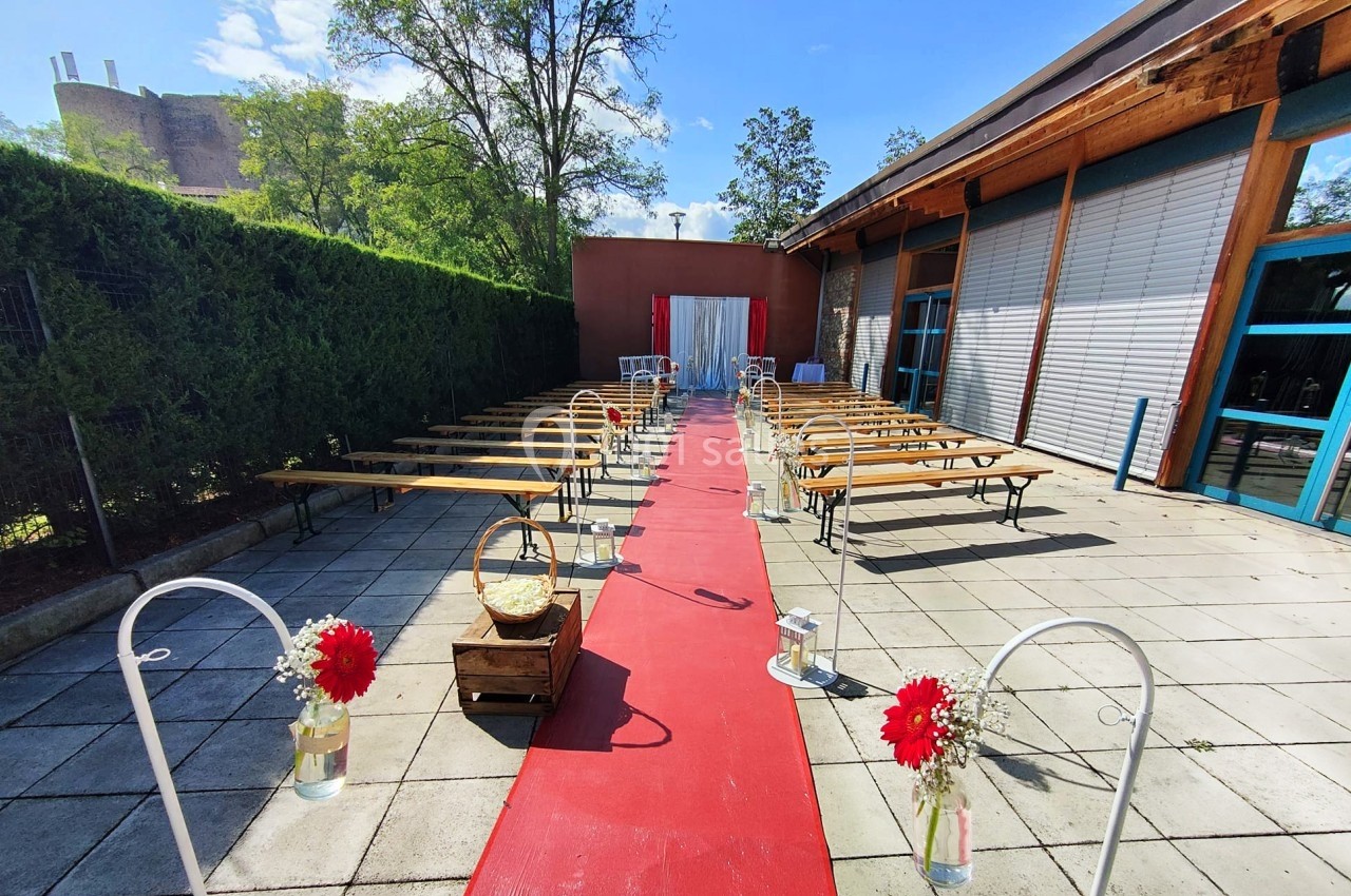 Allée centrale avec tapis rouge, bancs en bois de chaque côté, décorations florales et fond avec rideau blanc.