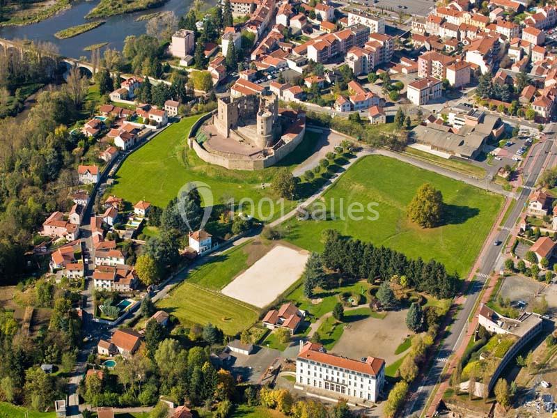 Vue aérienne d'un château médiéval entouré de verdure, au cœur d'une petite ville avec des maisons et des routes.