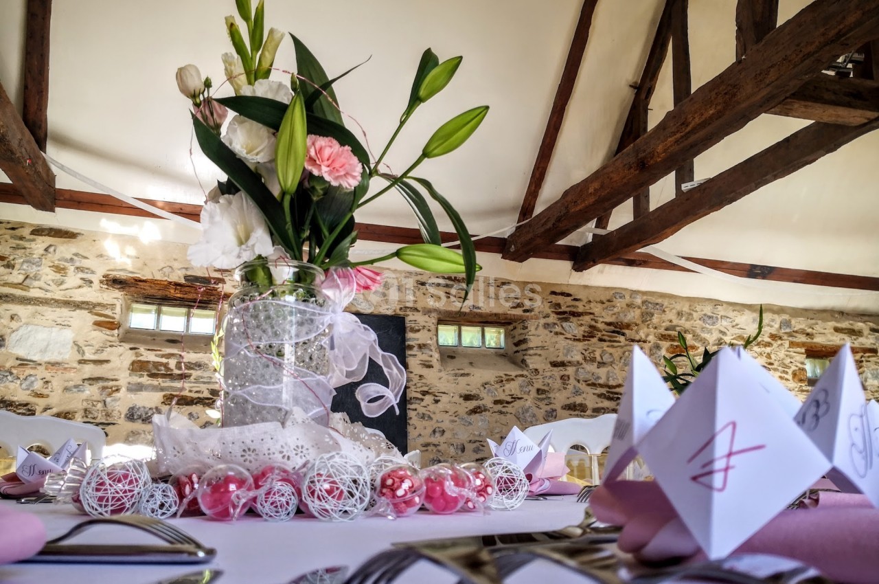 Centre de table floral avec des lys et des roses, décoré de rubans et perles, dans une salle aux poutres apparentes.