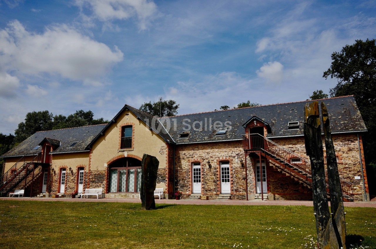 Bâtiment en pierre et crépi avec escaliers extérieurs, entouré de pelouse et d'arbres sous un ciel partiellement nuageux.