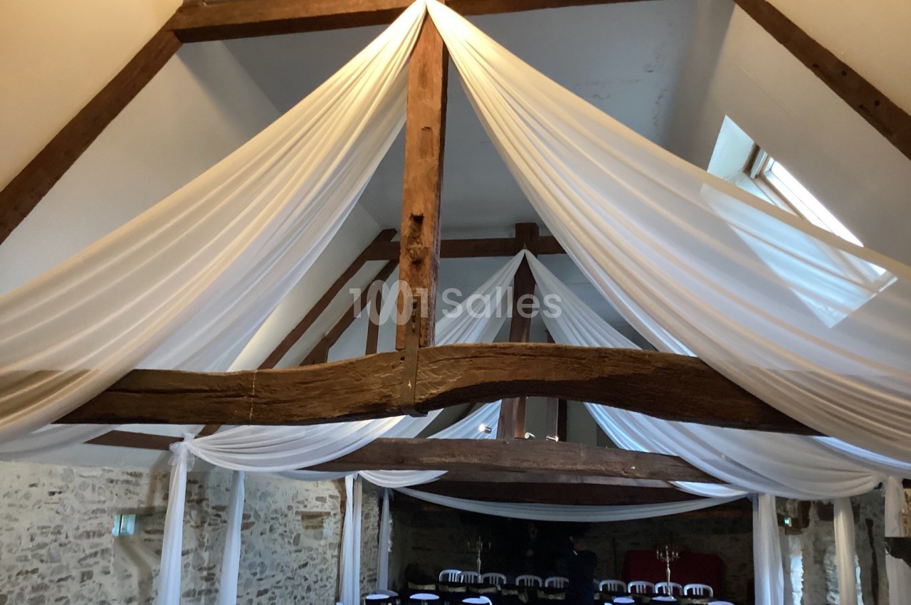 Plafond en poutres apparentes avec drapés blancs suspendus, dans une salle en pierre aménagée pour un événement.