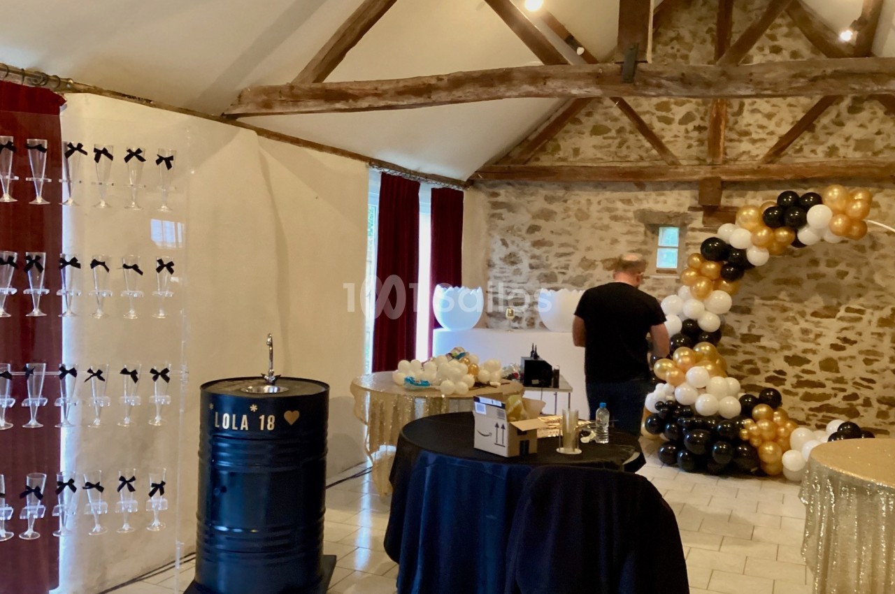 Salle décorée pour une fête avec ballons noirs, blancs et dorés, table ronde et présentoir à flûtes.