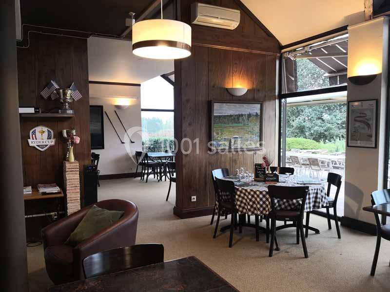 Salle de restaurant chaleureuse avec tables dressées, décor en bois, grandes fenêtres et vue sur un espace extérieur…