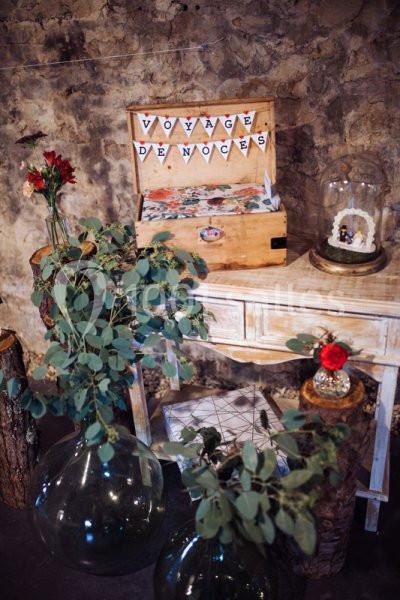 Décoration de mariage avec une boîte en bois ’Voyage de noces’, fleurs rouges et feuillage sur une table rustique.