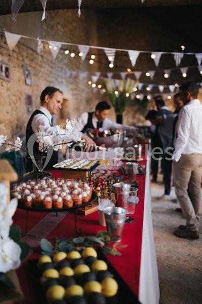 Table de buffet décorée avec des desserts, boissons et fleurs, dans une salle en pierre éclairée par des guirlandes…