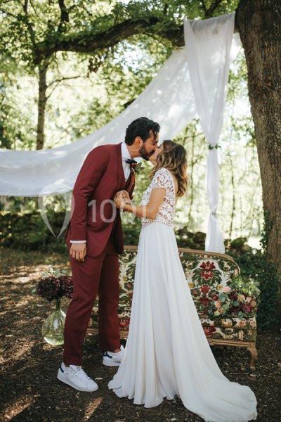 Un couple en tenue de mariage s'embrasse dans un décor extérieur avec des arbres et des drapés blancs.