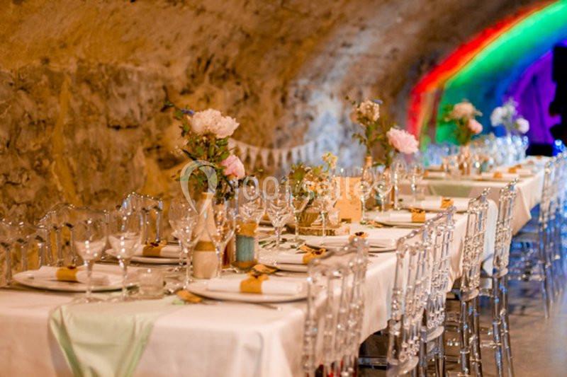 Tables décorées avec nappes blanches, fleurs et vaisselle élégante dans une salle voûtée éclairée par des lumières colorées.