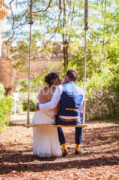 Un couple assis sur une balançoire en bois dans un jardin ensoleillé, entouré de verdure.