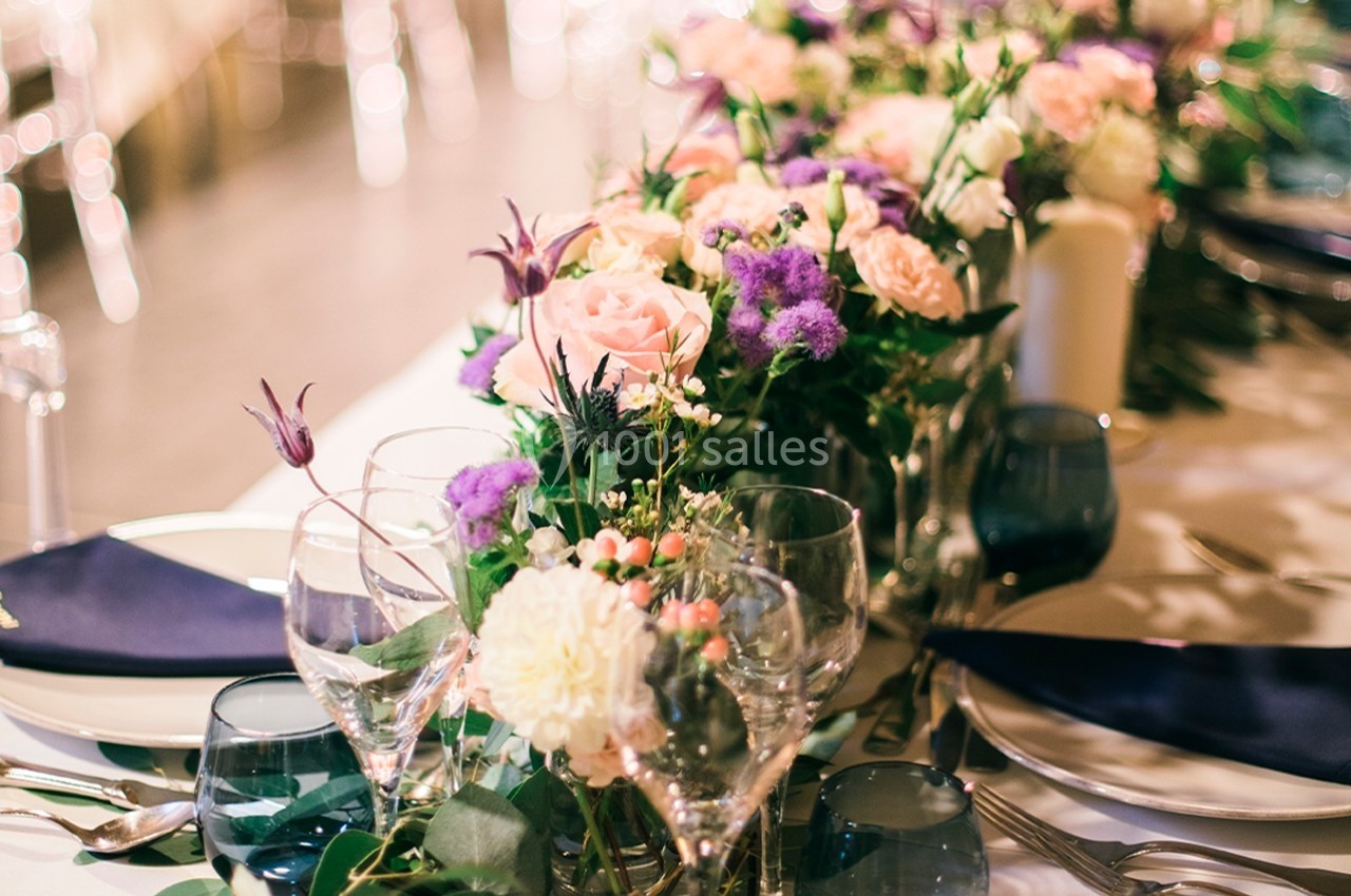Centre de table floral avec roses, fleurs violettes et feuillage, entouré de verres et assiettes sur une table dressée.