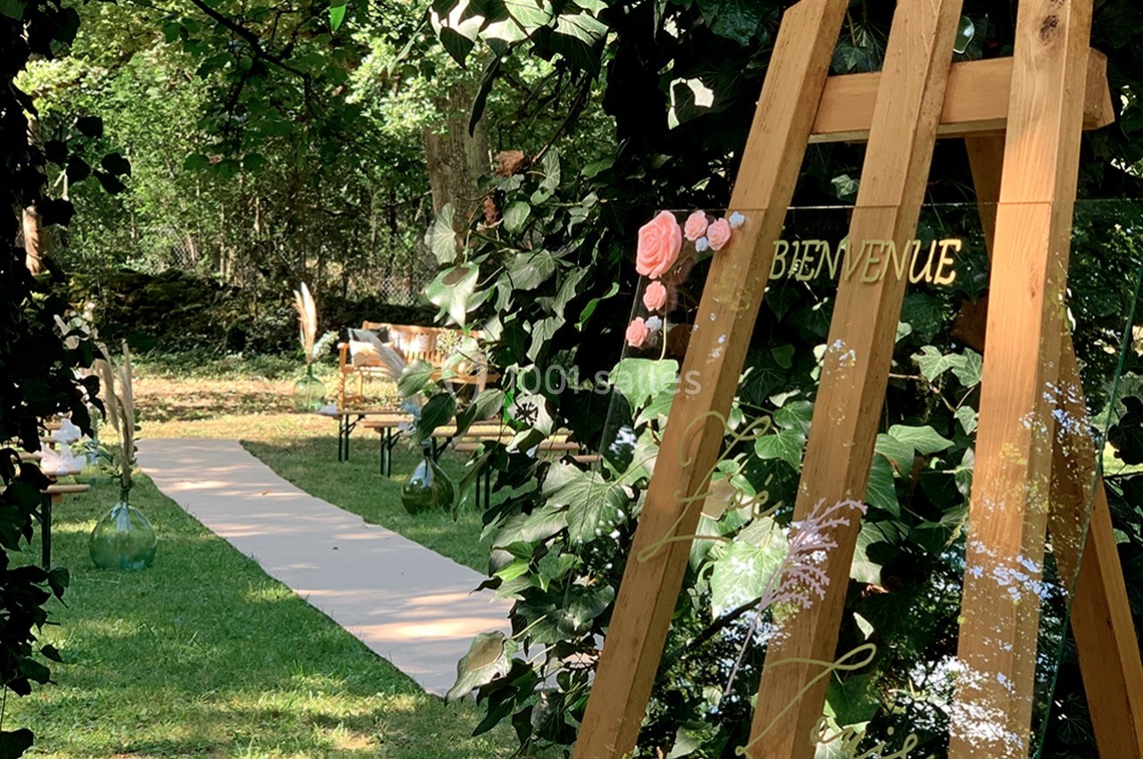 Chevalet en bois décoré de fleurs et d'un panneau ’Bienvenue’, placé dans un jardin verdoyant avec une allée aménagée.