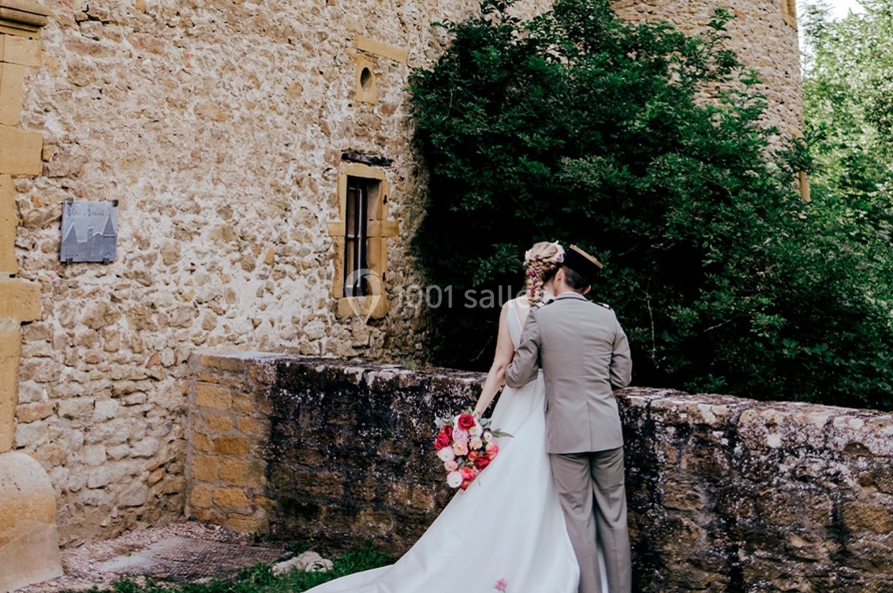 Un couple en tenue de mariage se tient près d'un mur en pierre, devant un bâtiment ancien entouré de verdure.